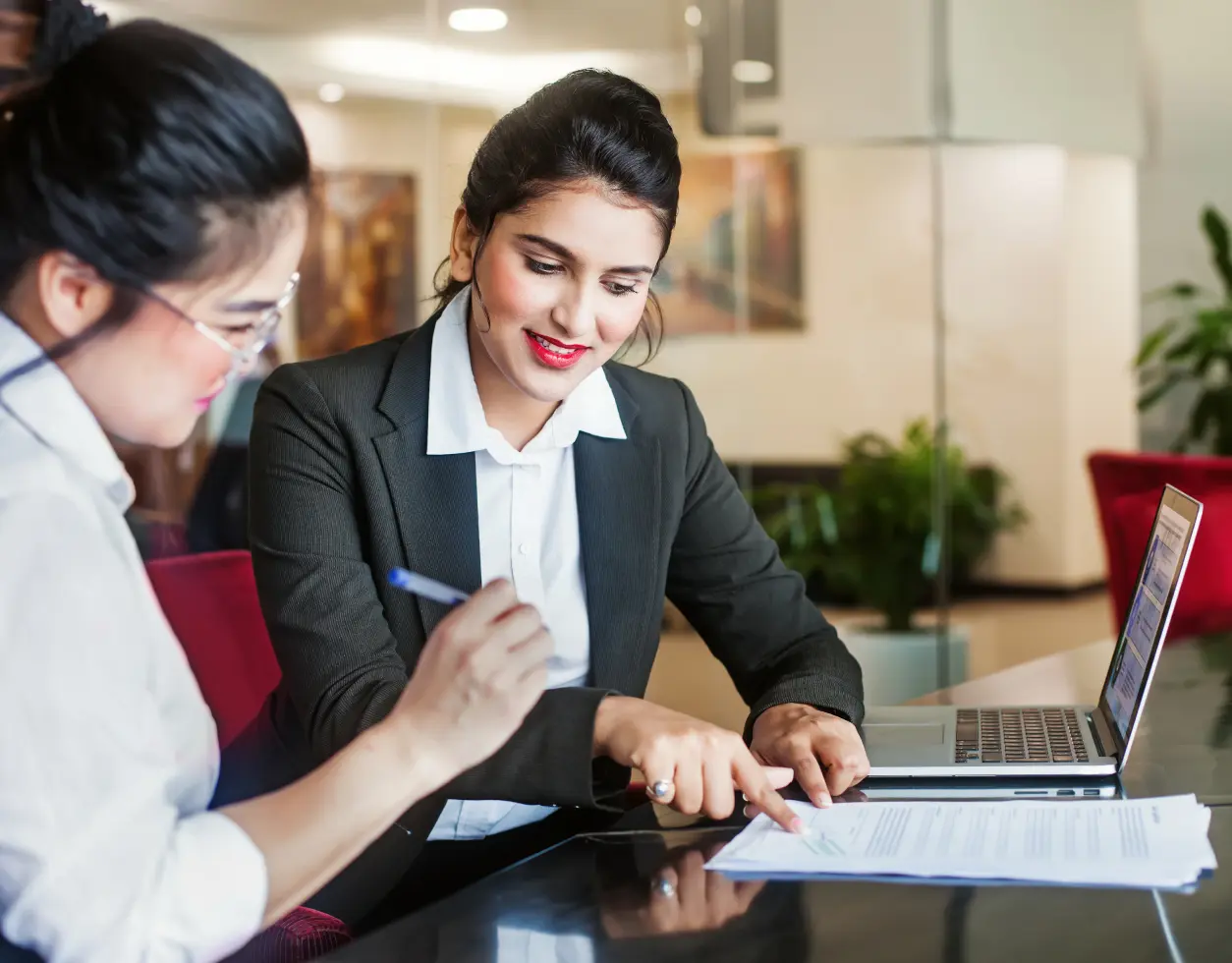 Two women are sitting at a table and one is looking at the paper.